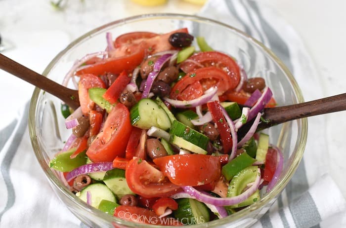 Tomato wedges, cucumber moons, bell pepper and onion slices tossed with vinaigrette in a large glass bowl with dark wooden spoons. 