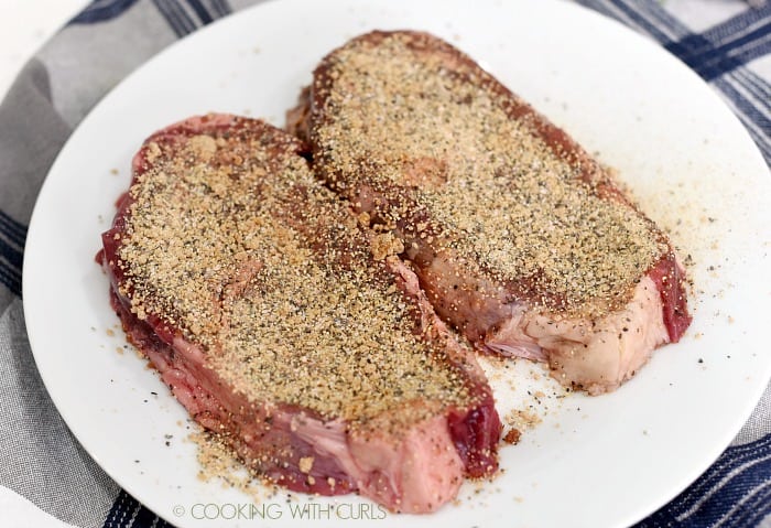 Two ribeye steaks coated with seasoning mixture laying on a white dinner plate. 