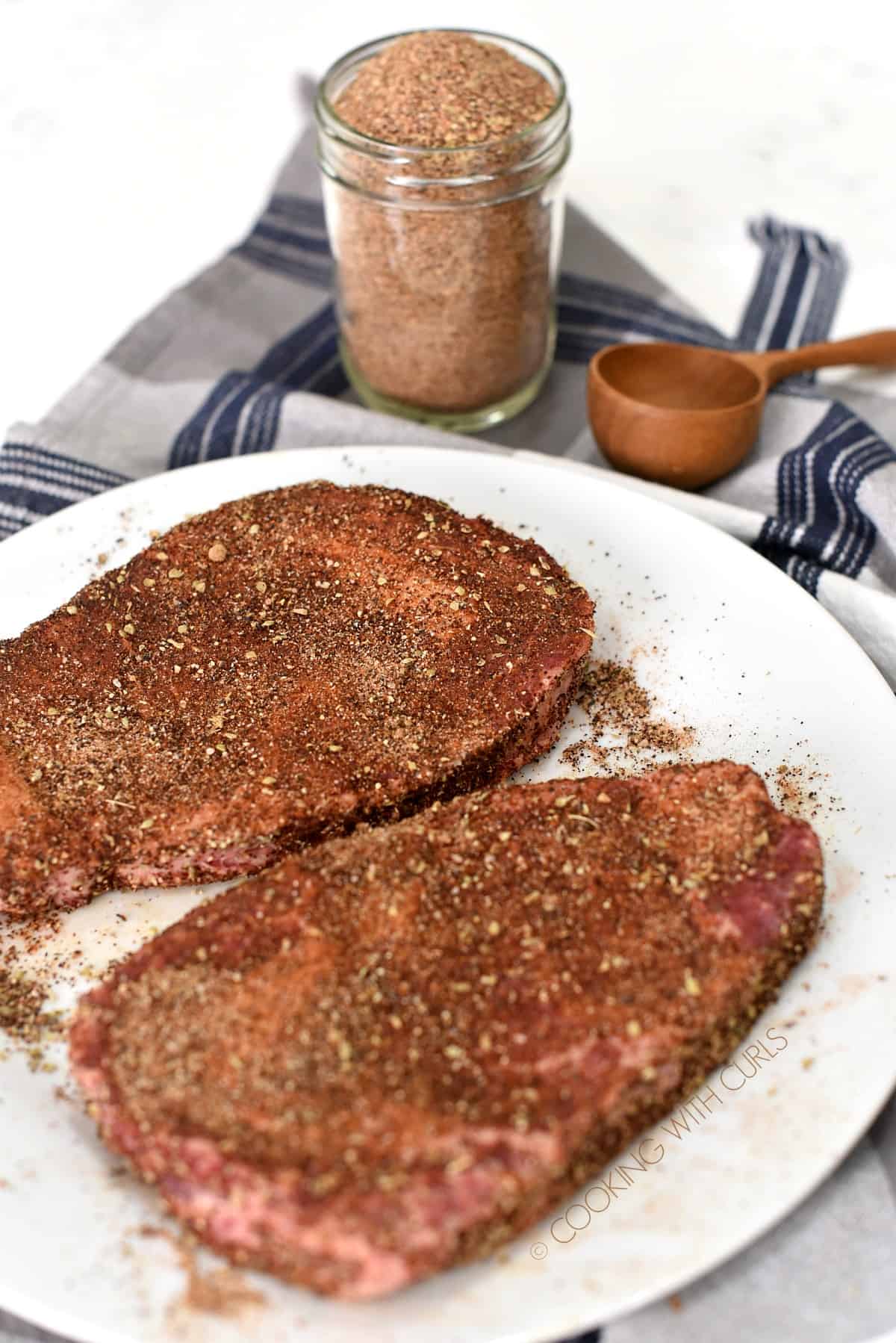 Two steaks rubbed with cowboy steak rub laying on a white plate with a jar of spice mix in the background.