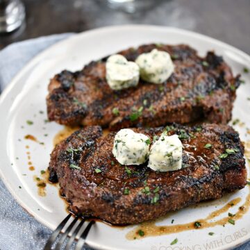 Two Perfect Grilled Steaks topped with two herb butter hearts sitting on a large white dinner plate.