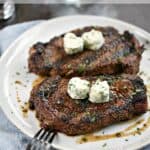 Two Perfect Grilled ribeye Steaks topped with two herb butter hearts sitting on a large white dinner plate.