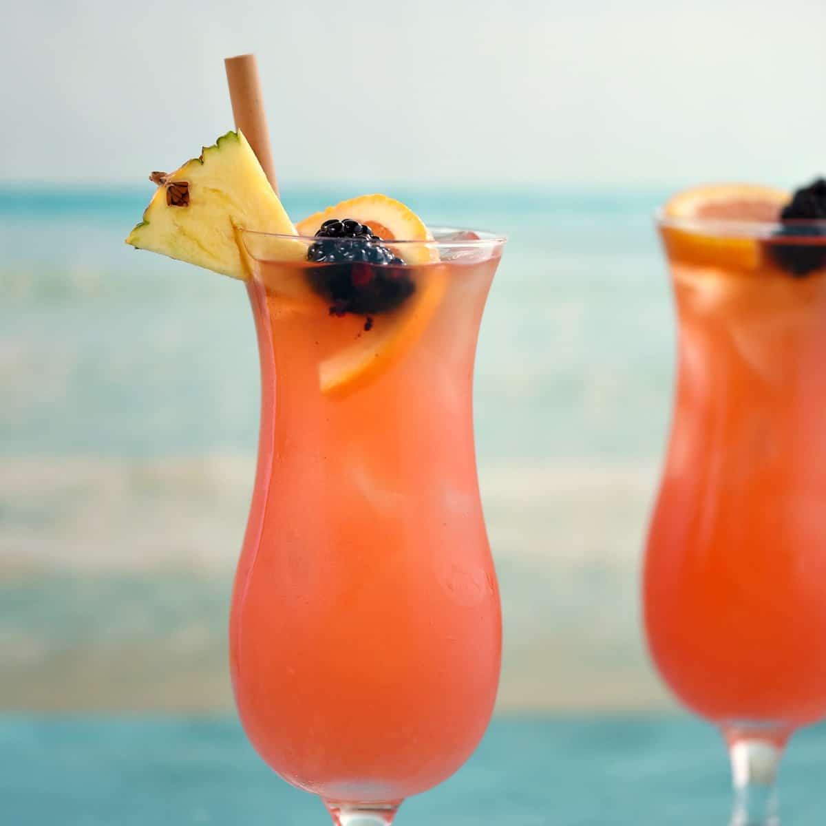 two hurricane glasses filled with a pink cocktail and garnished with a straw, pineapple wedge, orange slice and blackberry with an ocean image in the background.