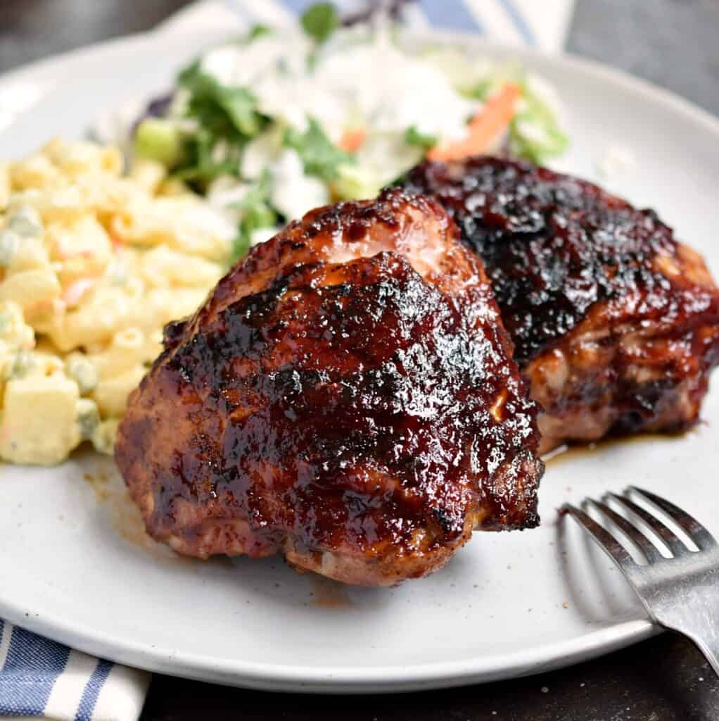 two barbecue chicken thighs on a white plate with Hawaiian potato salad and a green salad in the background.