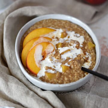 a big bowl of steel cut oats and chopped peaches topped with peach slices and heavy cream wrapped in a tan napkin with a black spoon sticking out of the bottom right side.