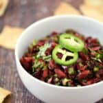 chopped cherries, jalapeno, and cilantro salsa in a white bowl topped with two jalapeno slices with pita chips scattered around the table.