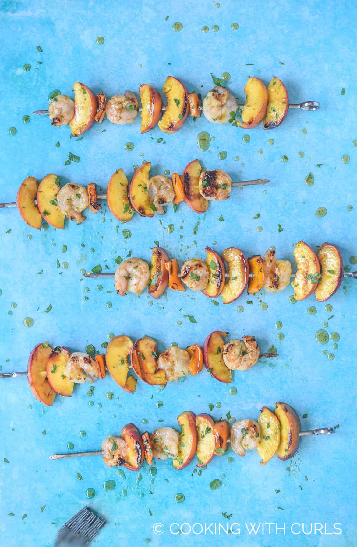 looking down on five metal skewers filled with shrimp, orange bell pepper and peach wedges drizzled with glaze on a blue background.