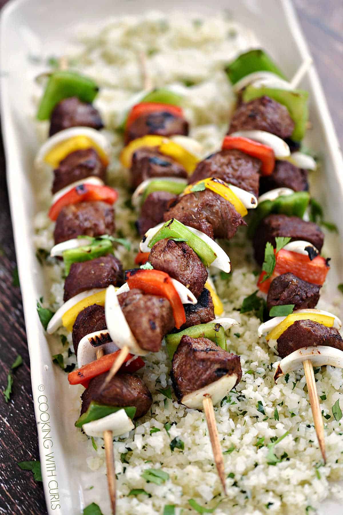 Four wooden skewers with grilled steak cubes, onion slices and bell pepper squares laying on a bed of cilantro lime rice.
