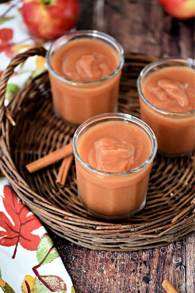 three jars filled with apple butter on a wicker tray with cinnamon sticks, a fall napkin and apples in the background.