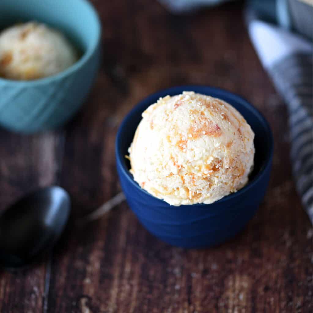 looking down on a small dark blue bowl with a scoop of peach ice cream sitting next to a black spoon, with a small turquoise bowl of ice cream in the upper left hand corner.