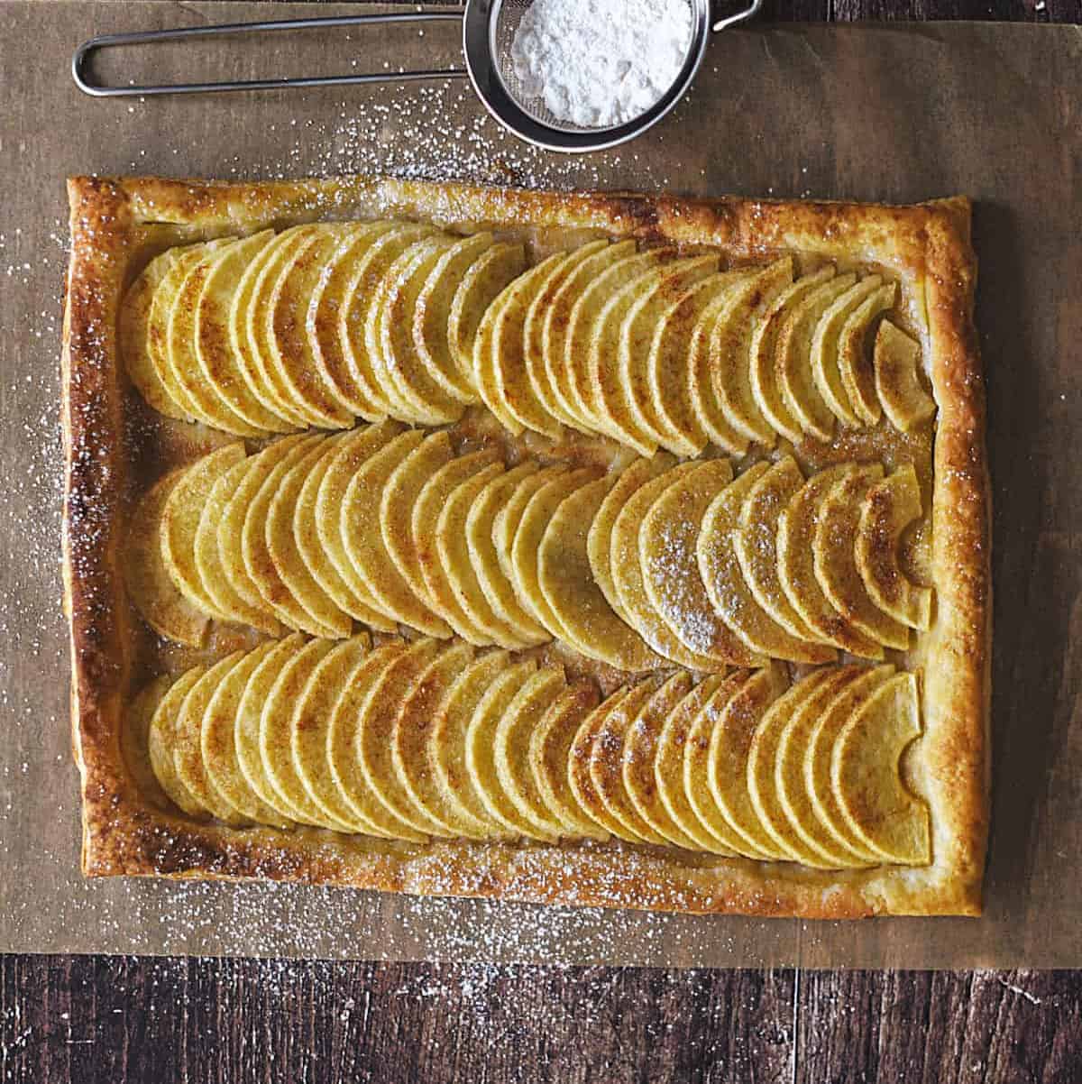 looking down on rows of apple slices on a puff pastry crust on a sheet of parchment paper with powdered sugar in a metal sifter at the top.