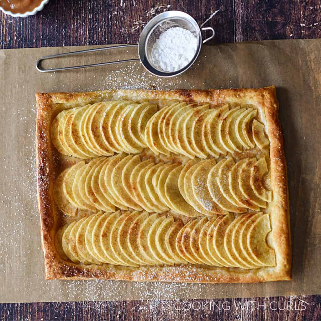 looking down on rows of apple slices on a puff pastry crust on a sheet of parchment paper with powdered sugar in a metal sifter at the top.