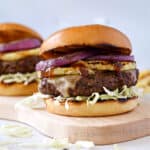 a wood cutting board with a big cheeseburger topped with grilled pineapple and red onion slices, shredded cabbage and teriyaki sauce on a brioche bun with a second burger in the background.