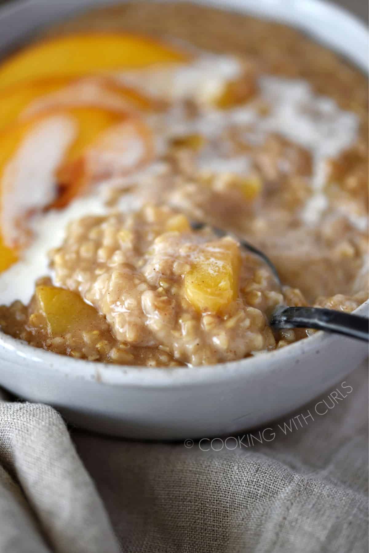 a close up image of steel cut oats and peaches on a black spoon being pulled out of the bowl of oatmeal.