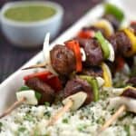 four wooden skewers with grilled steak cubes, onion slices and bell pepper squares laying on a bed of cilantro lime rice with a bowl of sauce on the side.