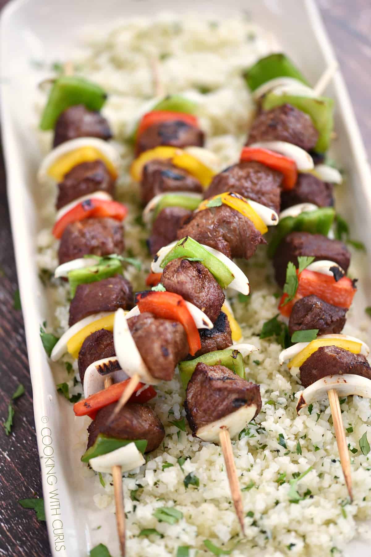 four wooden skewers with grilled steak cubes, onion slices and bell pepper squares laying on a bed of cilantro lime rice.