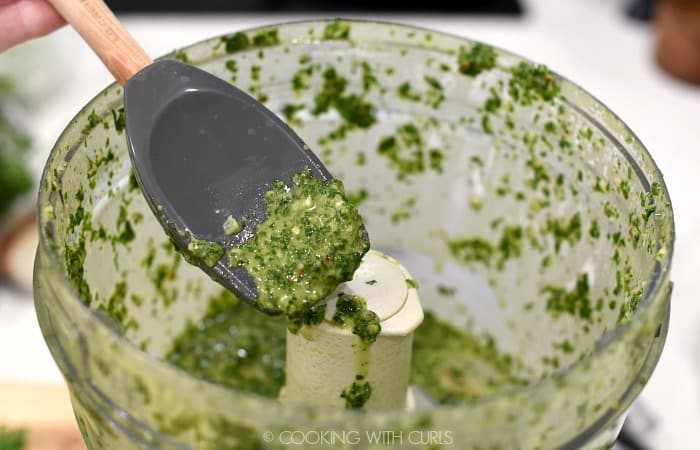 a gray silicone spatula holding a scoop of chimichurri over the bowl of a food processor. 