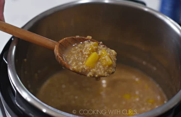 a wooden spoon holding cooked steel cut oats and chopped peaches over the pot of oatmeal. 
