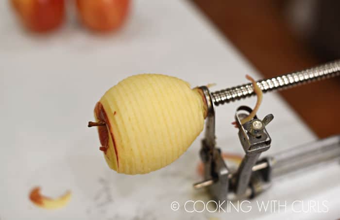 apple on the end of an apple, peeler corer contraption attached to the counter top with two apple in background. 