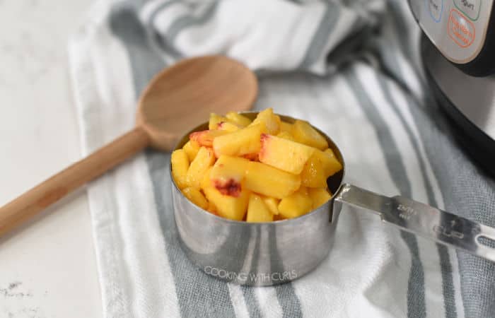 chopped peaches in a stainless steel measuring cup laying on a gray and white striped towel with a wooden spoon to the left and a pressure cooker to the right. 