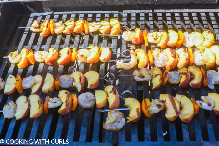 eight metal skewers with cooked shrimp, orange bell pepper and peach wedges on the grill. 