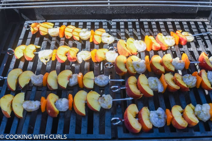 eight metal skewers with shrimp, orange bell pepper and peach wedges on the grill. 