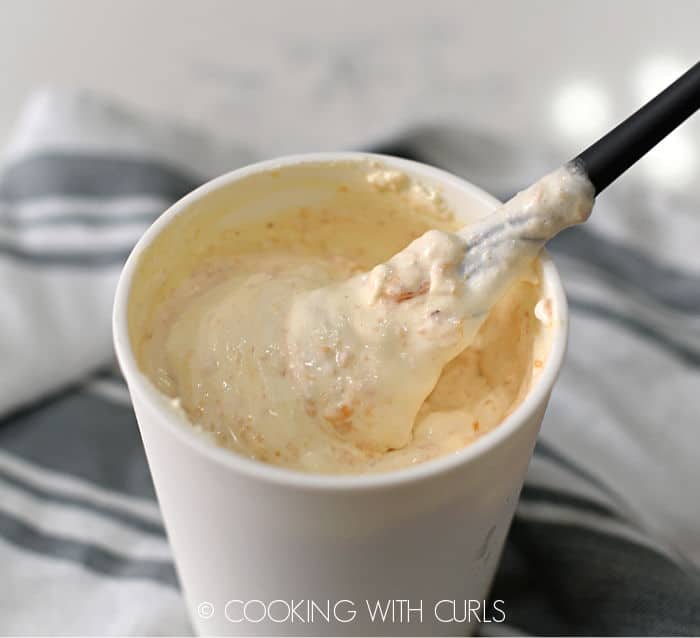 frozen peaches stirred into vanilla ice cream with a gray silicone spoon in a white plastic container sitting on a gray and white striped towel. 