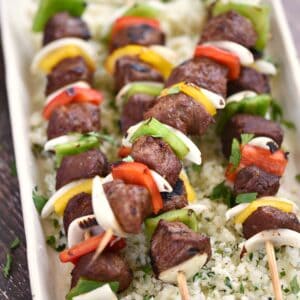 steak cubes, sliced onion, red, green and yellow peppers threaded on wooden skewers sitting on a bed of cilantro lime rice.