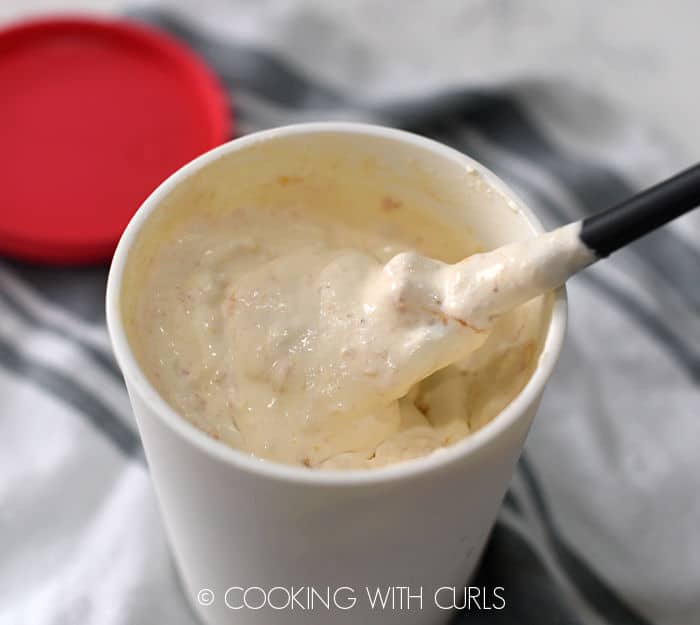 peach ice cream stirred with a gray silicone spoon inside a white plastic container with a red lid laying on a gray and white striped towel. 