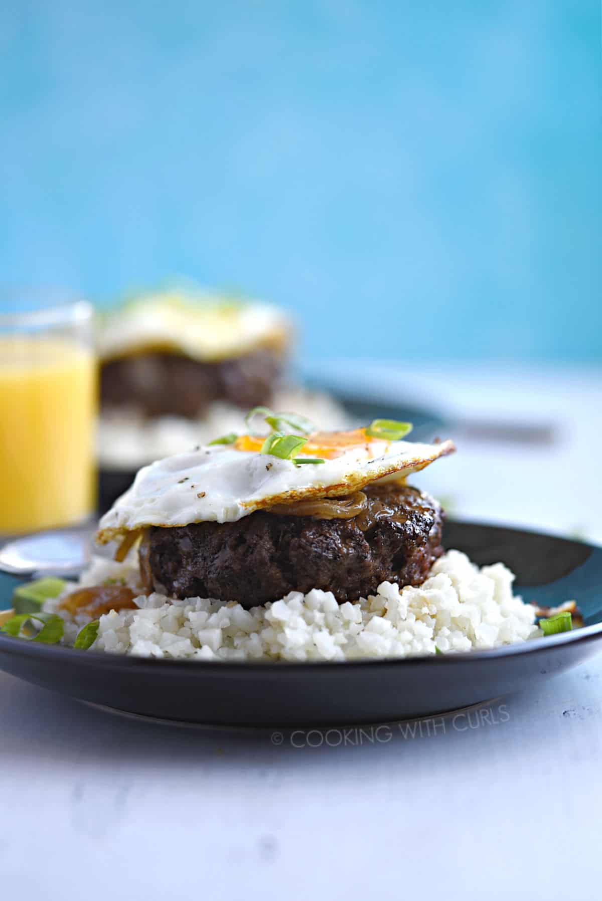 A ground hamburger steak patty topped with rich gravy and fried egg sits atop steamed rice and is garnished with green onions with a glass of orange juice and a second plate of food in the background.