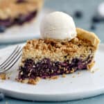 a slice of fresh blueberry crumb pie topped with ice cream on a white plate with a second pie in the background.