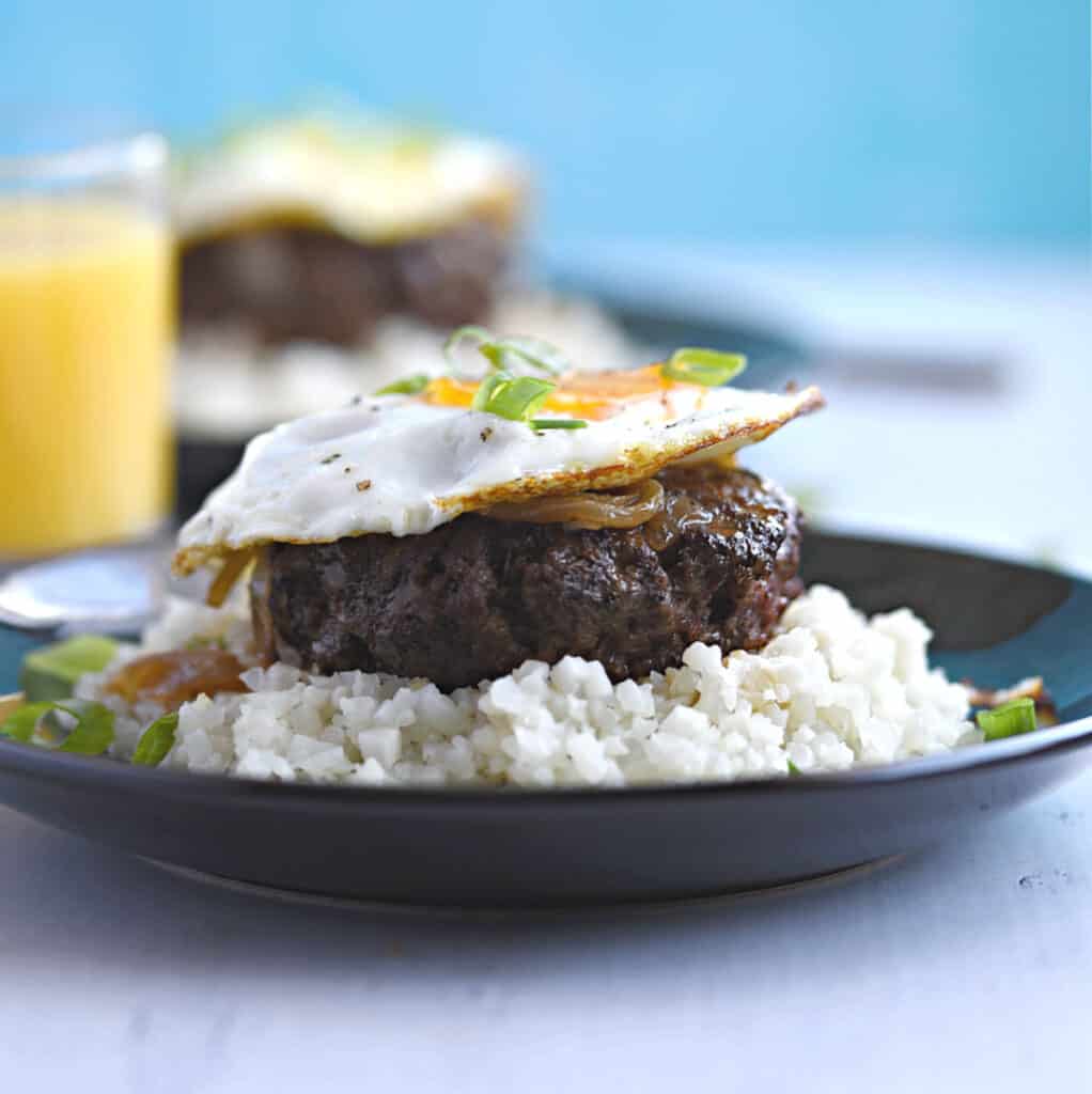 a steak burger patty topped with brown gravy, fried egg and green onions on a bed of rice with a glass of orange juice and a second plate of food in the background.
