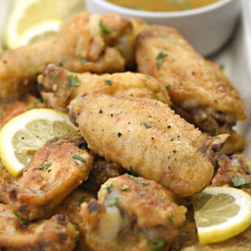 a pile of chicken wings on a white, rectangle platter with a small bowl of lemon garlic sauce in the right corner and lemon slices scattered over the wings.