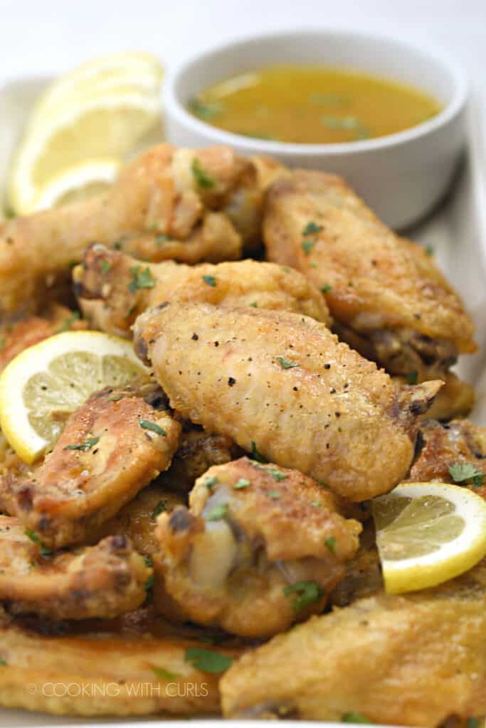 close up image of chicken wings piled up on a rectangle, white platter with a small bowl of lemon garlic sauce in the upper right hand corner and lemon slices scattered over the wings