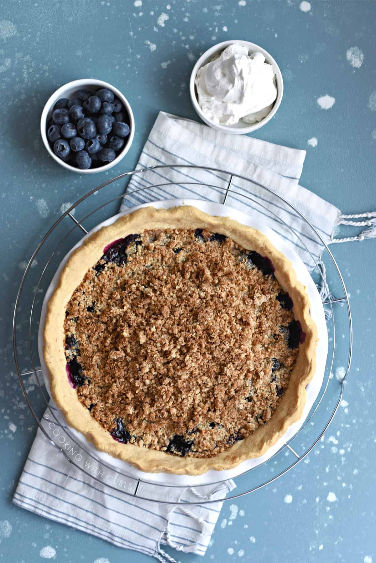 looking down on a crumb topped blueberry pie sitting on a wire cooling rack over a blue and white towel with a small bowl of blueberries and a bowl of whipped cream up above.