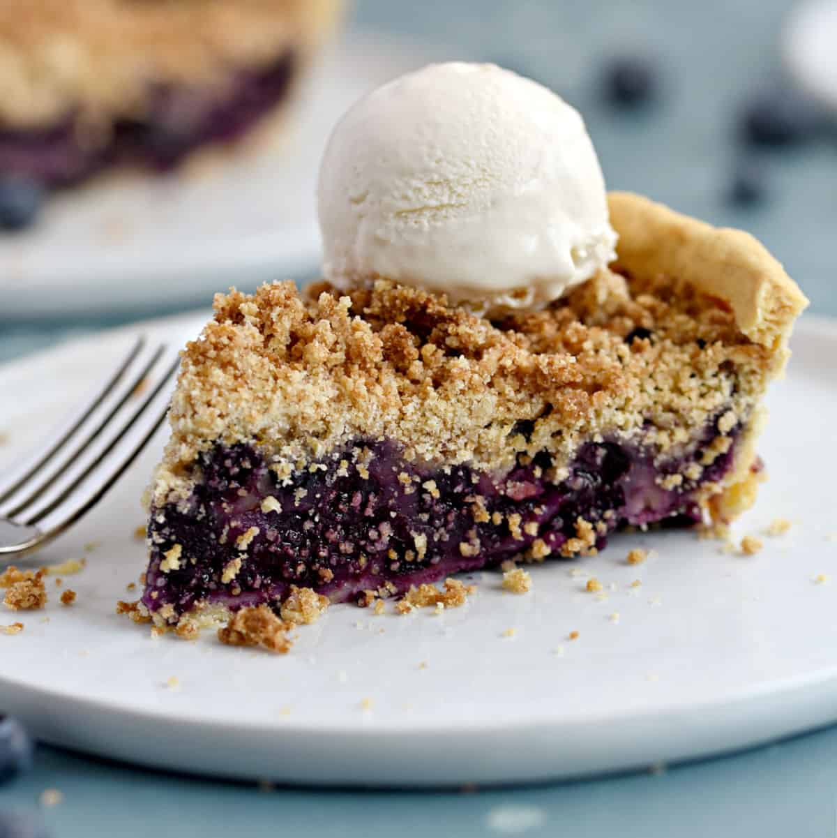 a slice of fresh blueberry crumb pie topped with ice cream on a white plate with a second pie in the background.