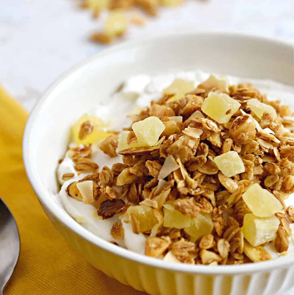 Pina Colada Granola topped yogurt in a white ribbed bowl sitting on a yellow napkin with a spoon to the left and spilled granola at the top.