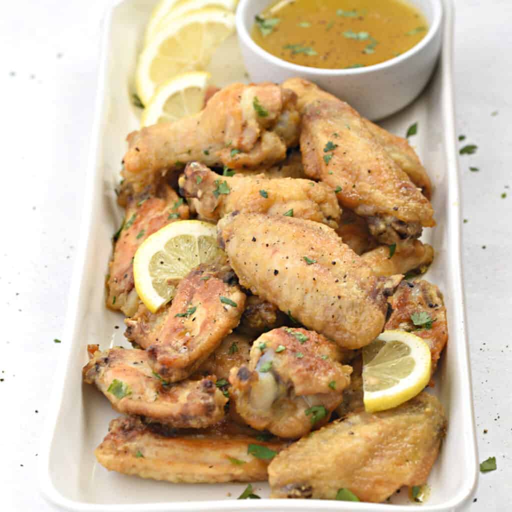 a close up image of a pile of chicken wings with a small bowl of lemon garlic sauce in the upper right hand corner.