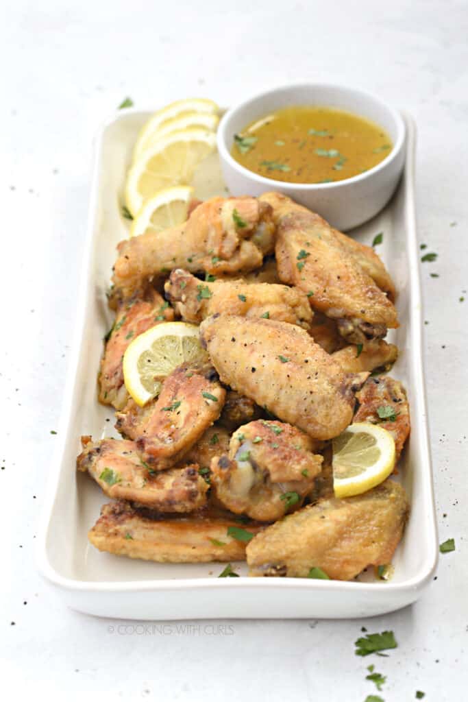 chicken wings piled up on a rectangle, white platter with a small bowl of lemon garlic sauce in the upper right hand corner and lemon slices scattered over the wings.