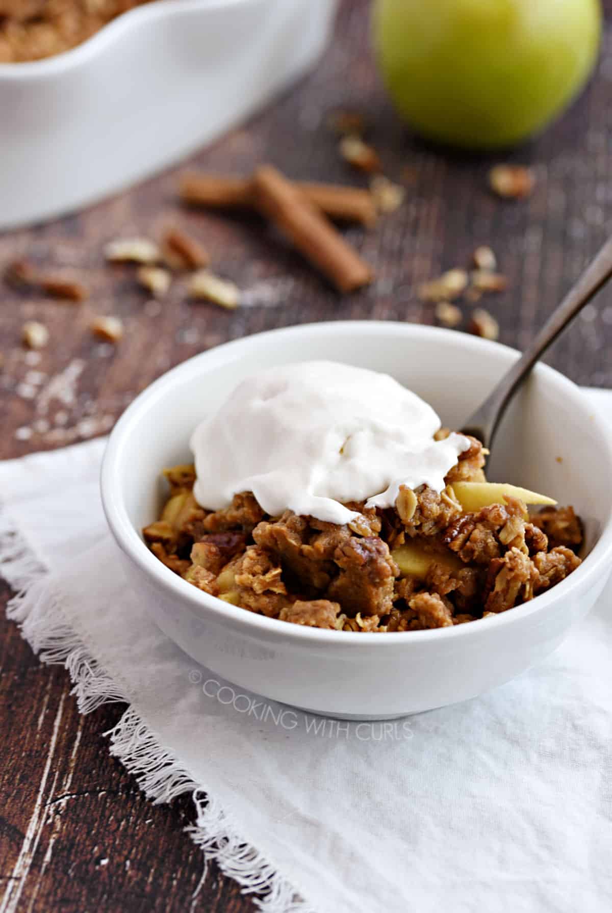 a white bowl with apple crisp topped with vanilla ice cream sitting on a white fringed napkin with an apple and baking dish in the background.