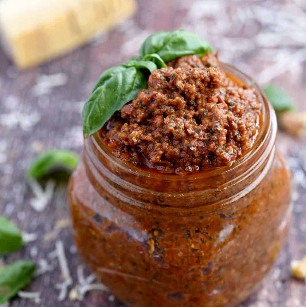 a square glass jar filled with sun-dried tomato pesto topped with fresh basil leaves.