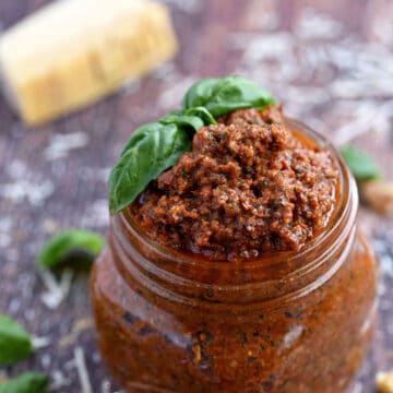 a square glass jar filled with sun-dried tomato pesto topped with fresh basil leaves.