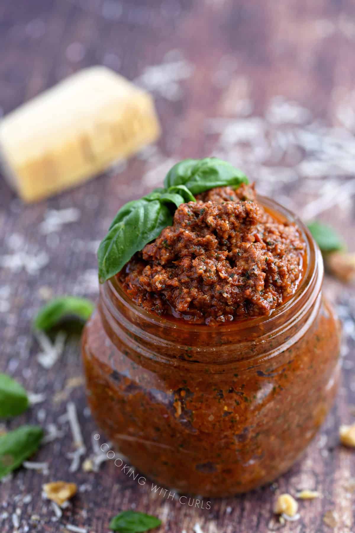 a square glass jar filled with sun-dried tomato pesto topped with fresh basil leaves.