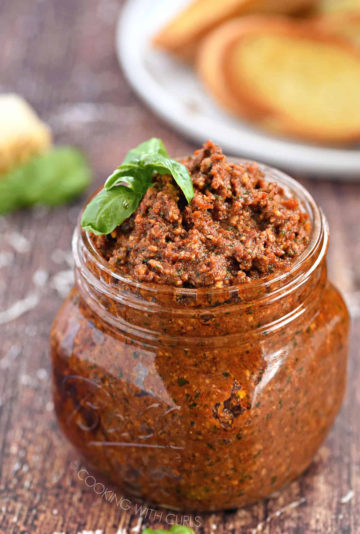 sun-dried tomato pesto in a glass jar with crostini and parmesan cheese in the background.