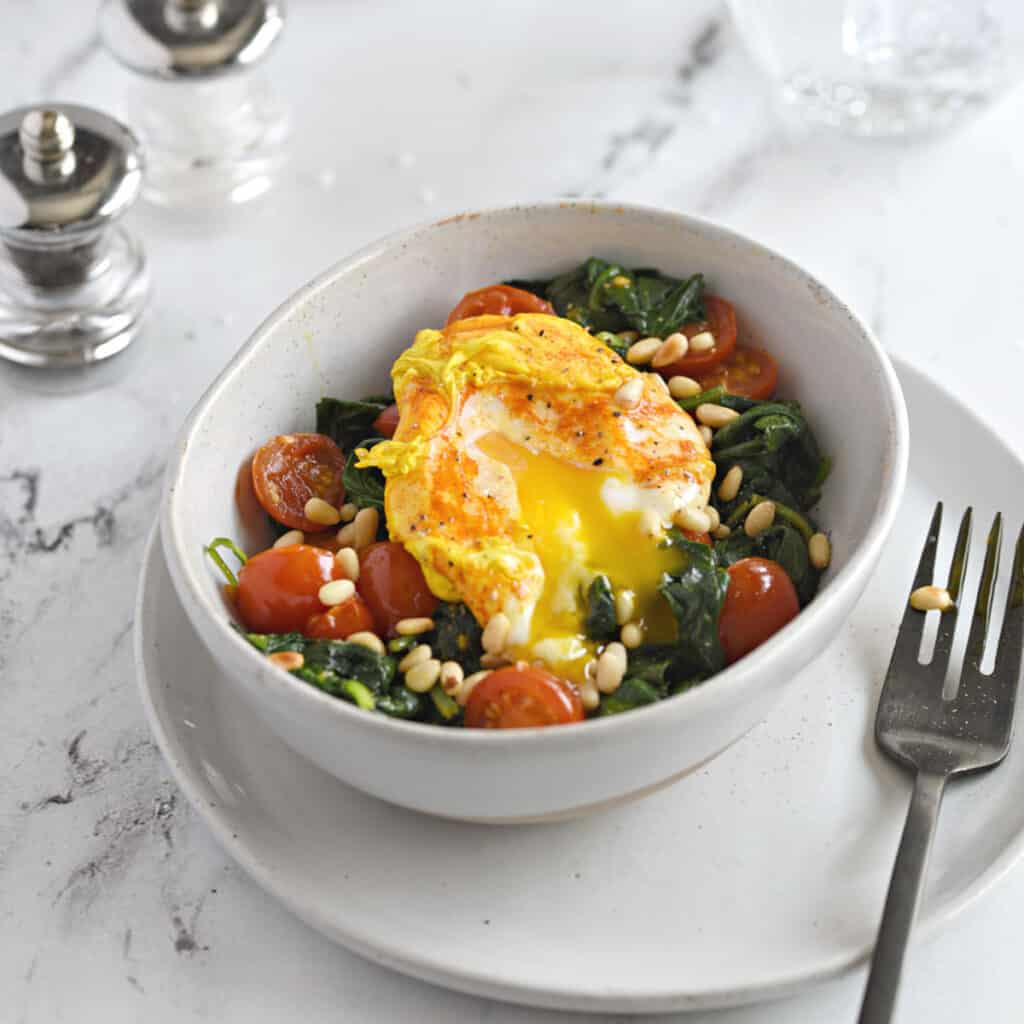 a turmeric poached egg split open over a bed of sauteed spinach and cherry tomato halves sprinkled with toasted pine nuts.