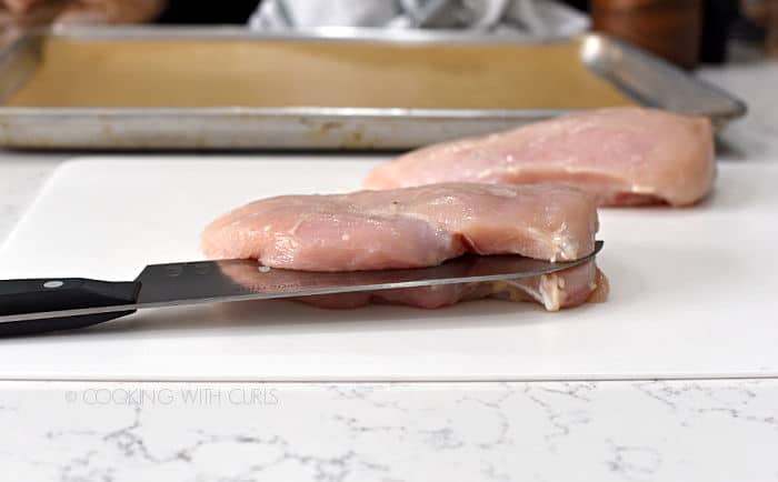 a chef knife slicing through the center of a chicken breast on a white cutting mat with a second breast in the background. 