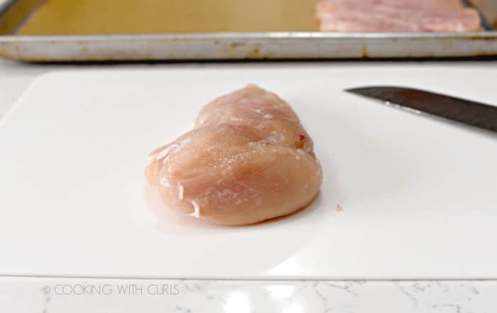 a raw chicken breast laying on a white plastic cutting mat with a chef knife on the right and a baking sheet with a butterflied chicken breast on a sheet of parchment paper. 