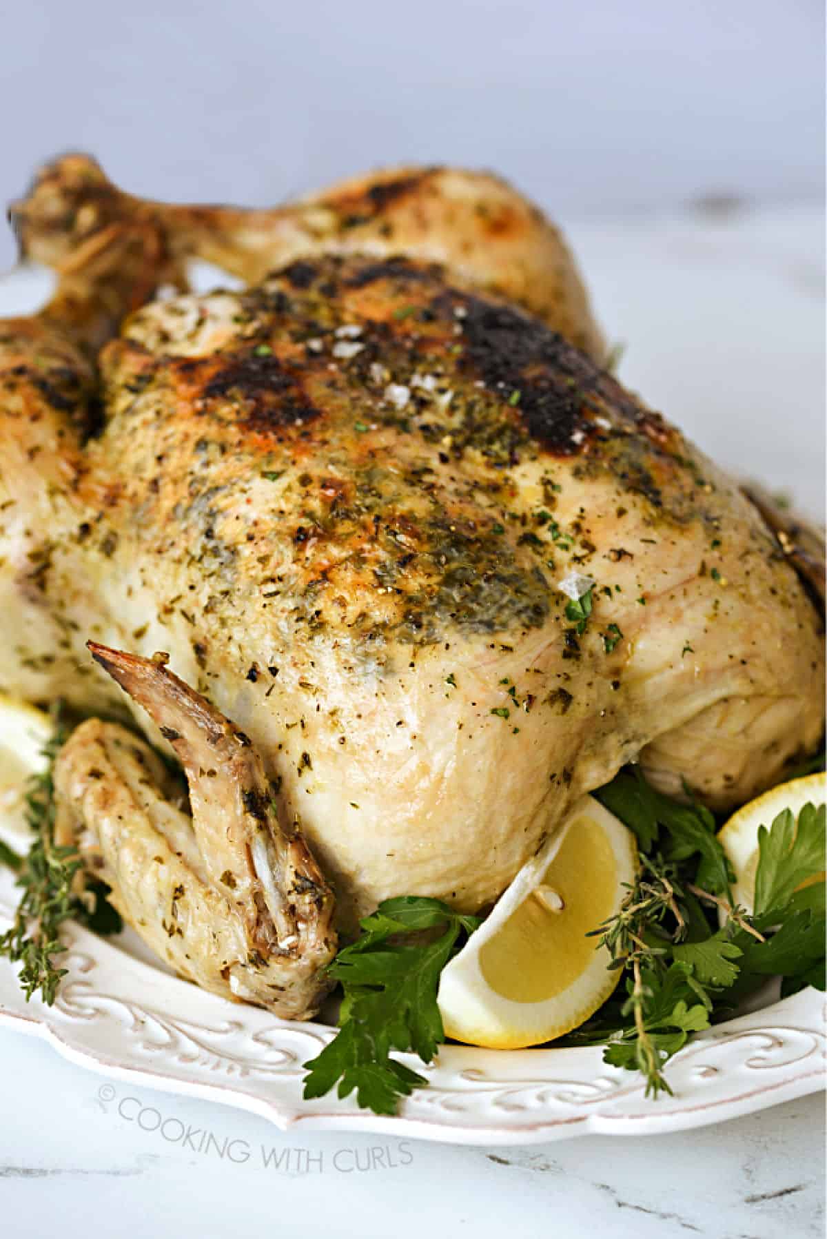 A close-up image of a whole chicken on a white platter surrounded by fresh herbs and lemon wedges.