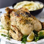 An Instant Pot Whole Greek Chicken on a white platter with thyme and lemon wedges, with a bowl of mashed potatoes in the background.