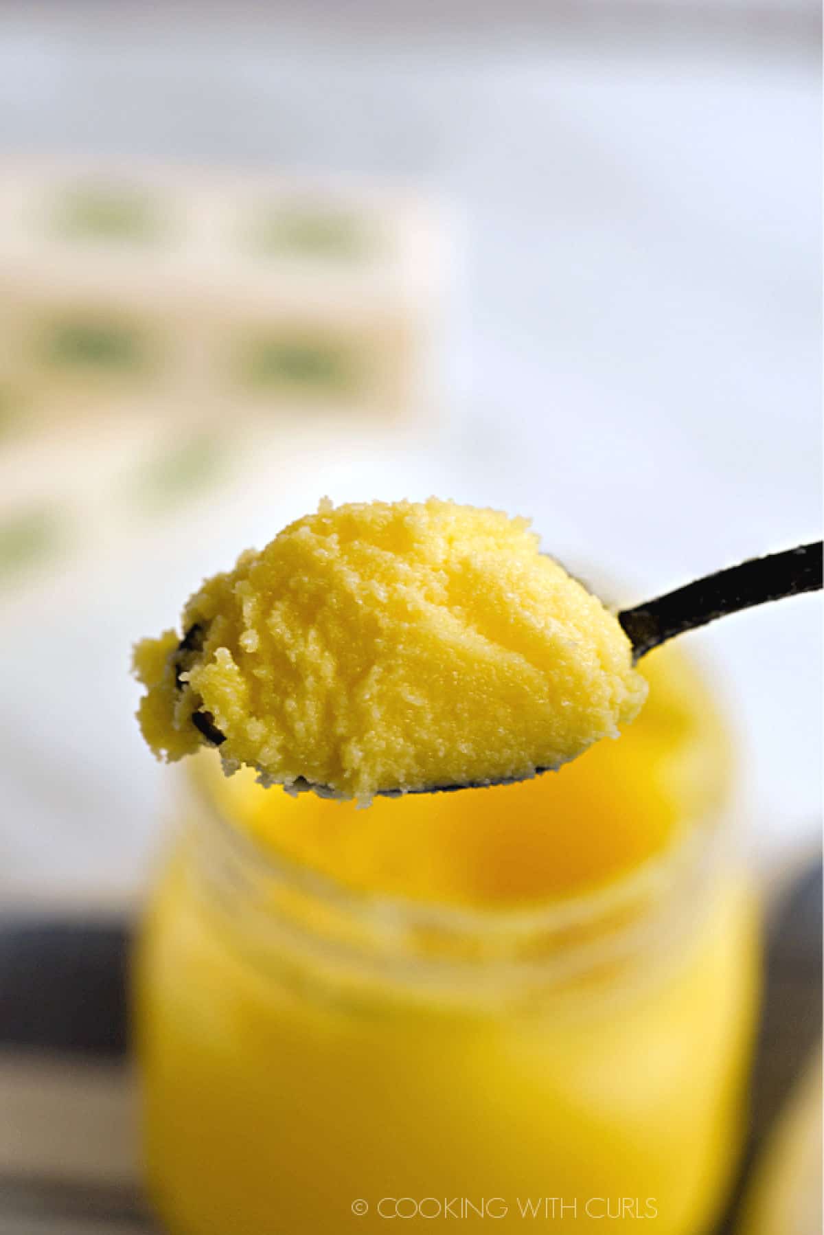 chilled ghee on a black spoon with a jar of ghee underneath and two sticks of butter in the background.