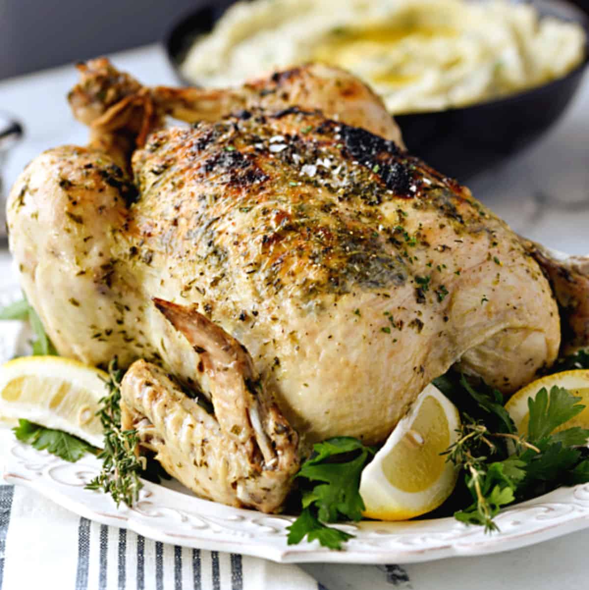 An Instant Pot Whole Greek Chicken on a white platter with a bowl of mashed potatoes in the background.